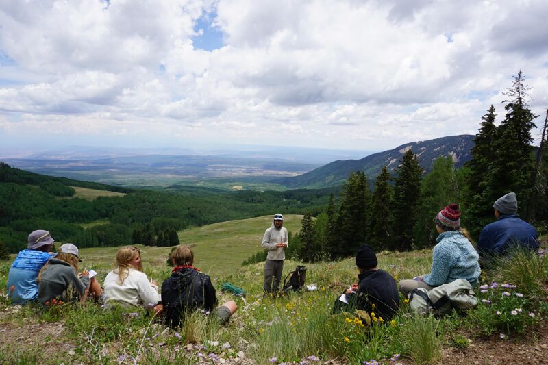 A group of people are gathered on a grassy hillside, seemingly listening to a person standing and gesturing towards the expansive valley and mountains in the background. The sky is filled with fluffy clouds, suggesting a pleasant day for an outdoor gathering. The scene conveys a sense of learning or discussion amidst a scenic natural environment.